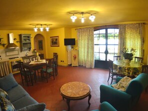 Ein Wohnzimmer mit einer Couch, einem runden Couchtisch, einem Esstisch mit Stühlen, einer Küchenzeile mit Herd und einem großen Fenster mit Vorhängen.