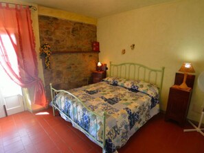 Ein Schlafzimmer mit einem Bett, einem Nachttisch, einer Lampe, einer Wand mit Steinoptik und einem Fenster mit Vorhang.