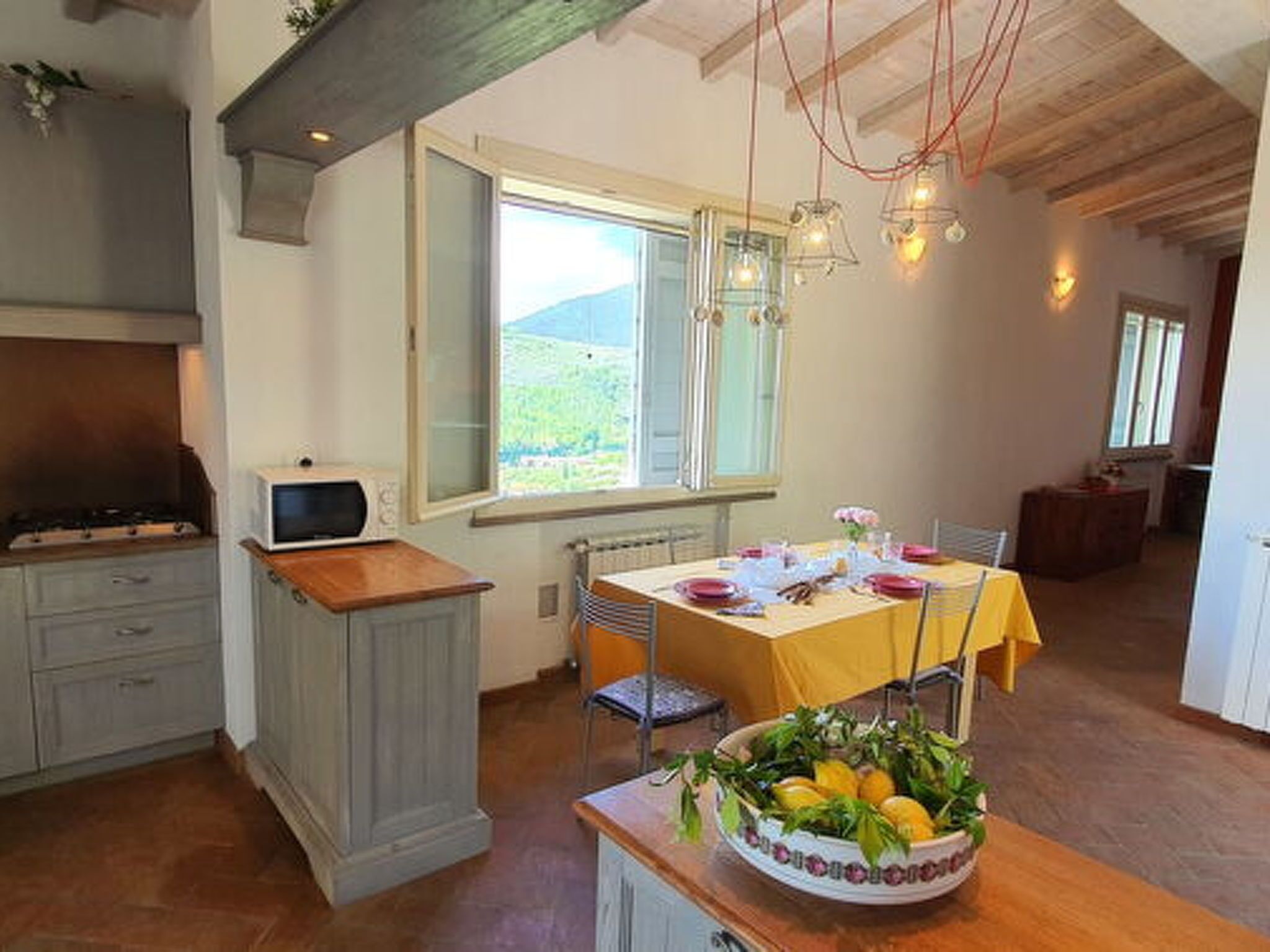 Una cocina con una mesa de comedor para dos personas, un frutero en la encimera y vistas al exterior a través de una ventana.