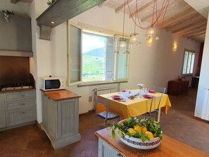 Een keuken met een eettafel voor twee personen, een fruitschaal op het aanrecht en een uitzicht op de natuur door een raam.