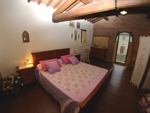 A bedroom with a wooden ceiling, a bed with floral bedding, a nightstand with a lamp, a dresser, and a window with curtains. - Le Case Di Carla Mela by Interhome (Borgo a Buggiano)