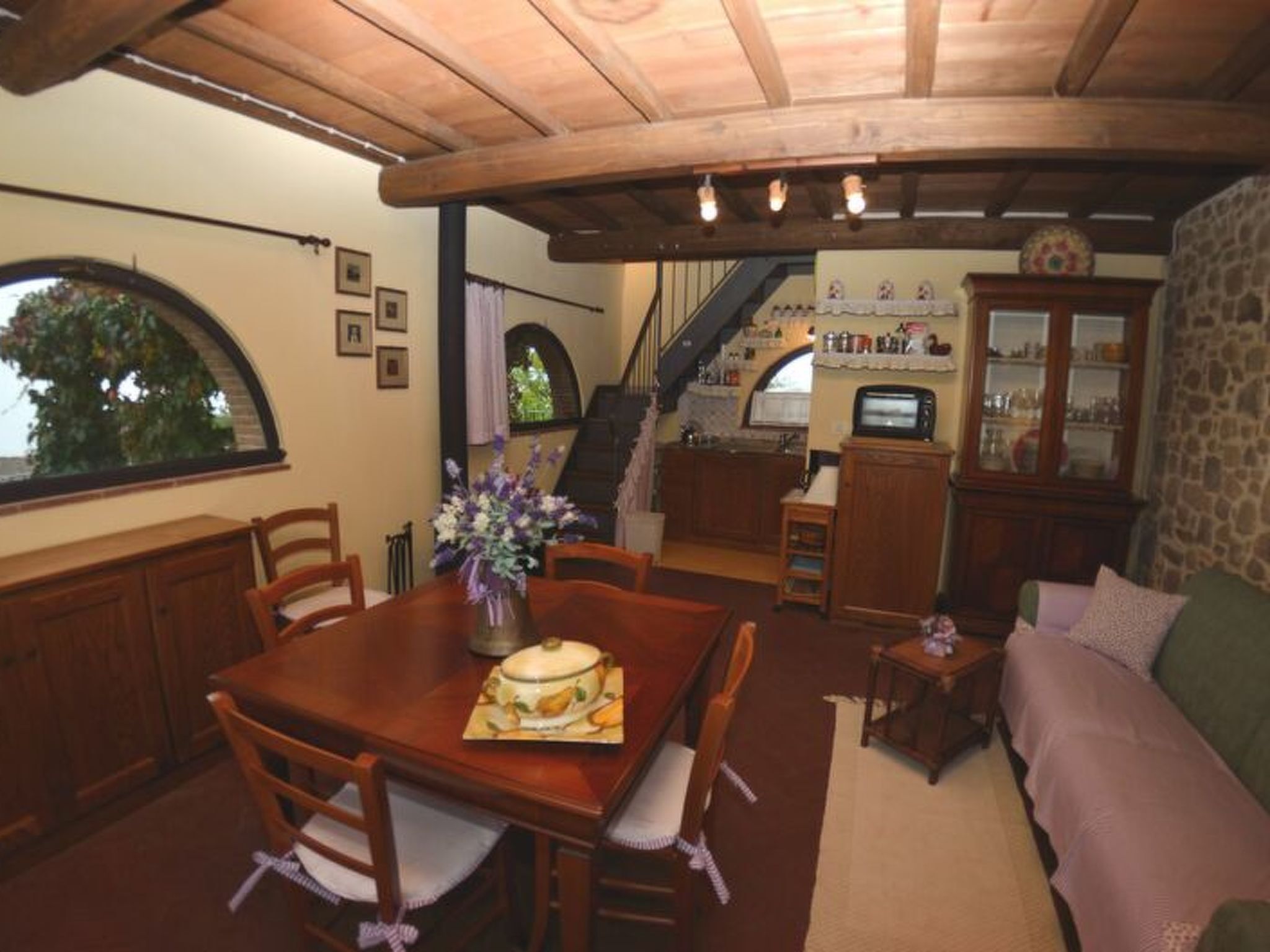 Zona pranzo con tavolo e sedie in legno, un vaso con fiori, una cucina sullo sfondo e una scala che conduce a un piano superiore.