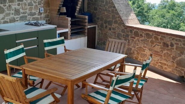 En udendørs terrasse med et træbord og stole, stenvægge og udsigt til grøn vegetation.