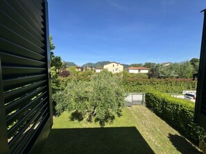 A view through a window with horizontal blinds, a grassy yard, a tree, and houses in the background. - Dependance Egle by Interhome (Ciciana)
