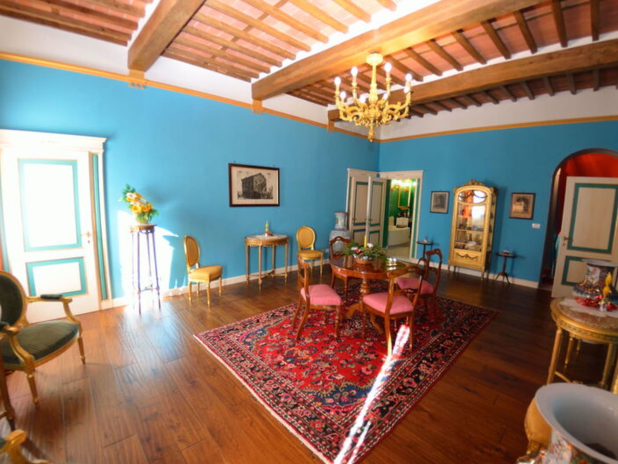 A room with a wooden ceiling, a chandelier, a dining table with chairs, a patterned rug, and a mirror.