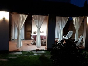 Ein überdachter Außenbereich mit Tisch und Stühlen, verhangen mit weißen Vorhängen, und ein kleiner Garten mit Grünpflanzen.