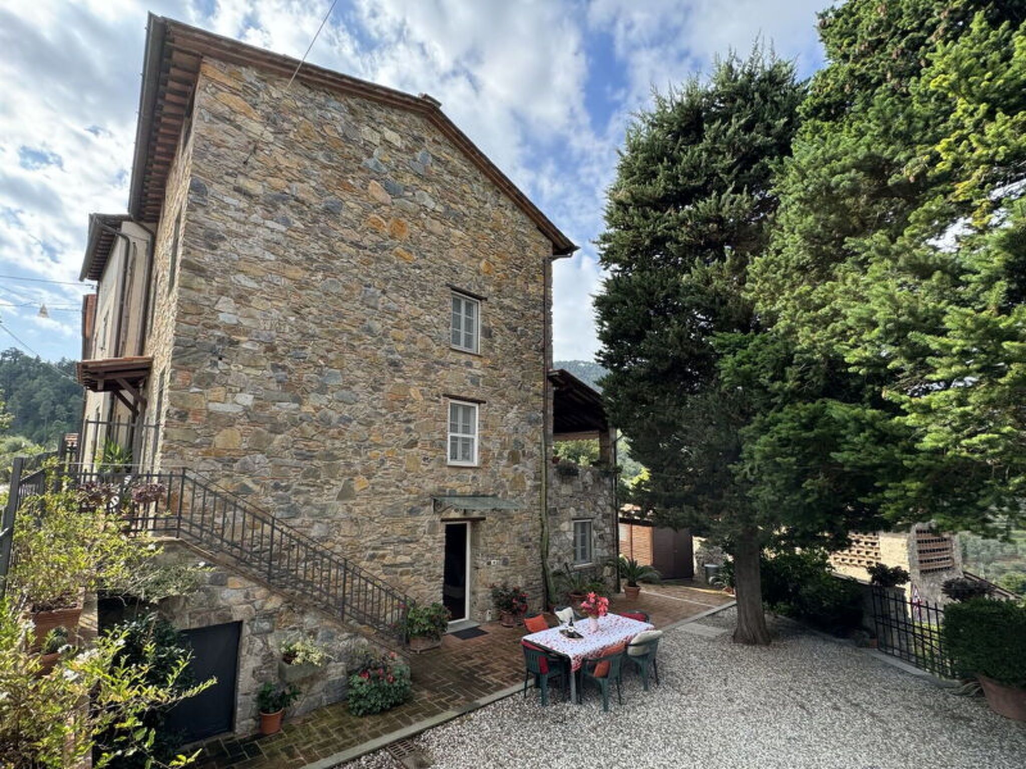 A stone building with a staircase, a patio with a table and chairs, and a large tree.