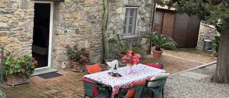 Un bâtiment en pierre avec une table et des chaises sur une terrasse, entouré de plantes en pot et d’un arbre.