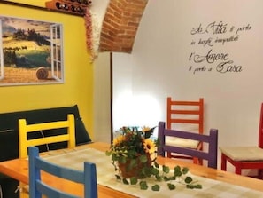 A dining area with a table set for two, two chairs, a vase with flowers, and a wall with a scenic painting and a decorative phrase. - Bocelli by Interhome (Peccioli)