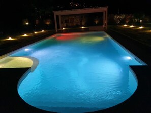 Ein Schwimmbecken bei Nacht mit beleuchteten Rändern und einem überdachten Bereich im Hintergrund.