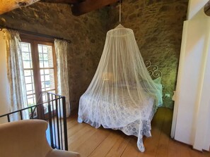 Una habitación con una cama bajo dosel blanco, suelo de madera, ventana con cortinas y pared de piedra.