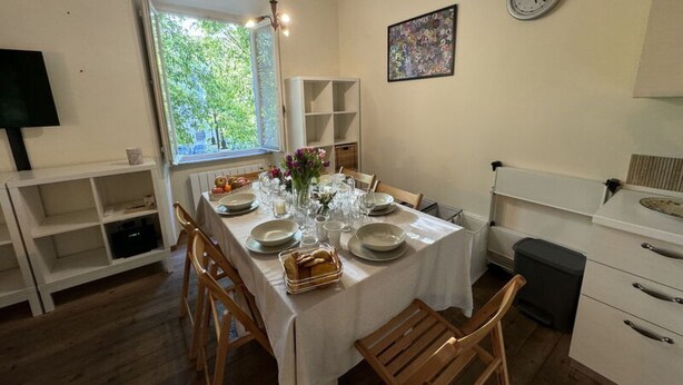 Un comedor con una mesa lista para cuatro personas, sillas de madera, un ventanal con vistas a la vegetación, un reloj en la pared y una estantería con libros.
