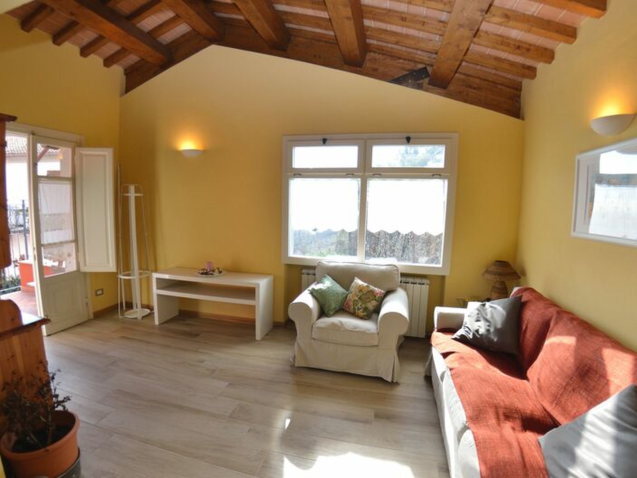 Un salon avec un plafond haut, des poutres en bois, un canapé blanc, un canapé rouge, un fauteuil blanc et une fenêtre donnant sur un beau paysage.