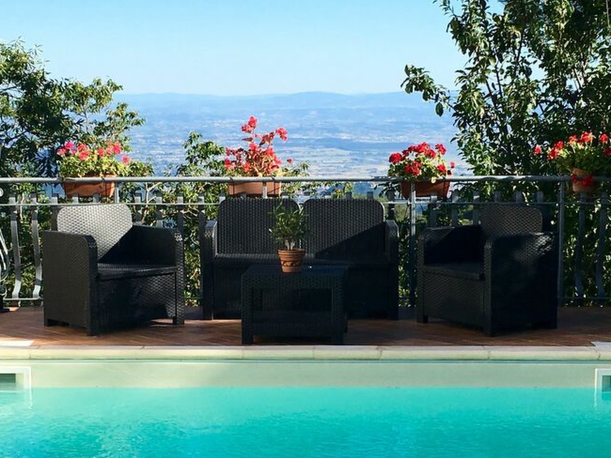A poolside area with wicker furniture and potted plants, overlooking a scenic view.