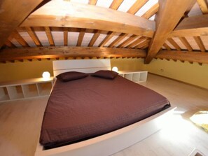 Un lit simple dans une chambre dont le plafond est incliné et orné de poutres en bois, avec une tête de lit blanche et une table de chevet sur laquelle se trouve une lampe.