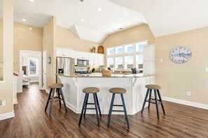 A kitchen with a central island, bar stools, and a clock on the wall. - Seabreeze at Ocean Hill: New to Rental Program, Private Pool & Hot Tub, Dog-Friendly (Corolla)
