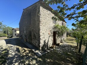 Ein Steinhaus mit kleinem Eingang, umgeben von einer Schotterauffahrt und Bäumen.