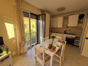 Uma sala de jantar com mesa posta para quatro pessoas, uma cozinha com armários e eletrodomésticos, e uma porta de vidro deslizante que dá para uma área externa.