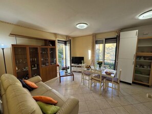 Ein Wohnzimmer mit Sofa, Esstisch, Stühlen, Fernseher und Schrank.