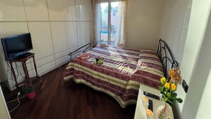 Une chambre à coucher avec un lit, un téléviseur posé sur un meuble, une fenêtre avec des rideaux et une plante en pot.