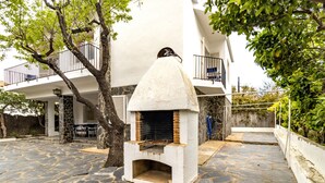 A stone patio with a fireplace, a tree, and a white building with a balcony. - 110 Port de la Selva - House close to the beach (Port De La Selva, El)