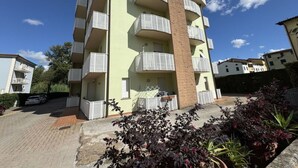 Um edifício residencial de vários andares, com varandas, entrada pavimentada e vegetação ao redor.