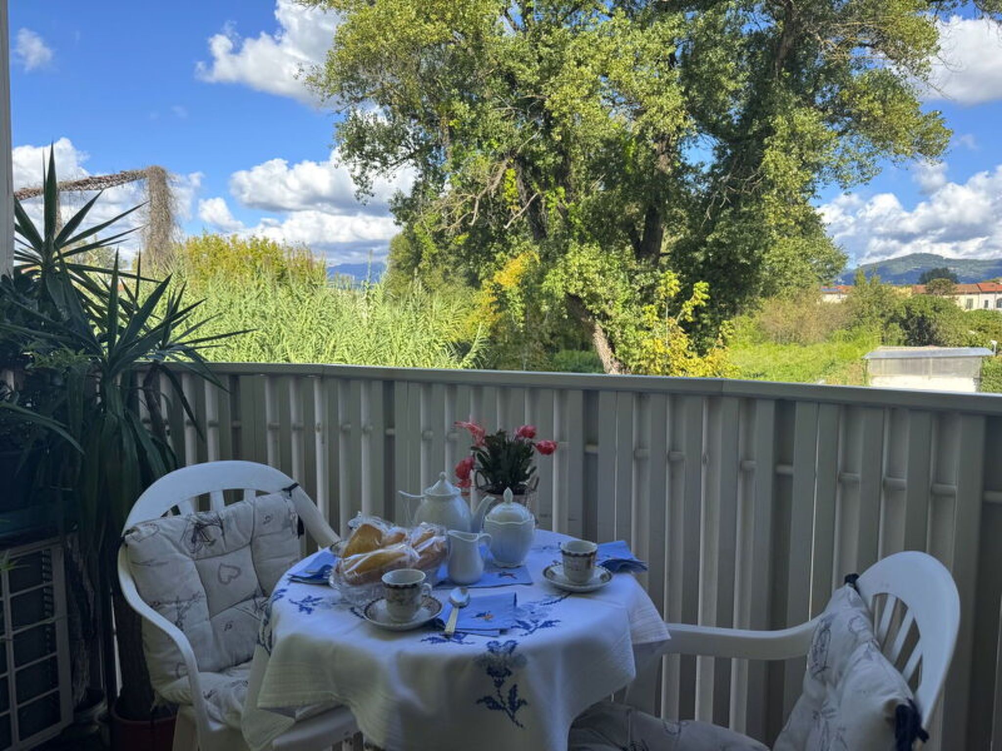 Un balcone con un tavolo apparecchiato per il tè, circondato da sedie bianche, con vista su alberi ed edifici in lontananza.