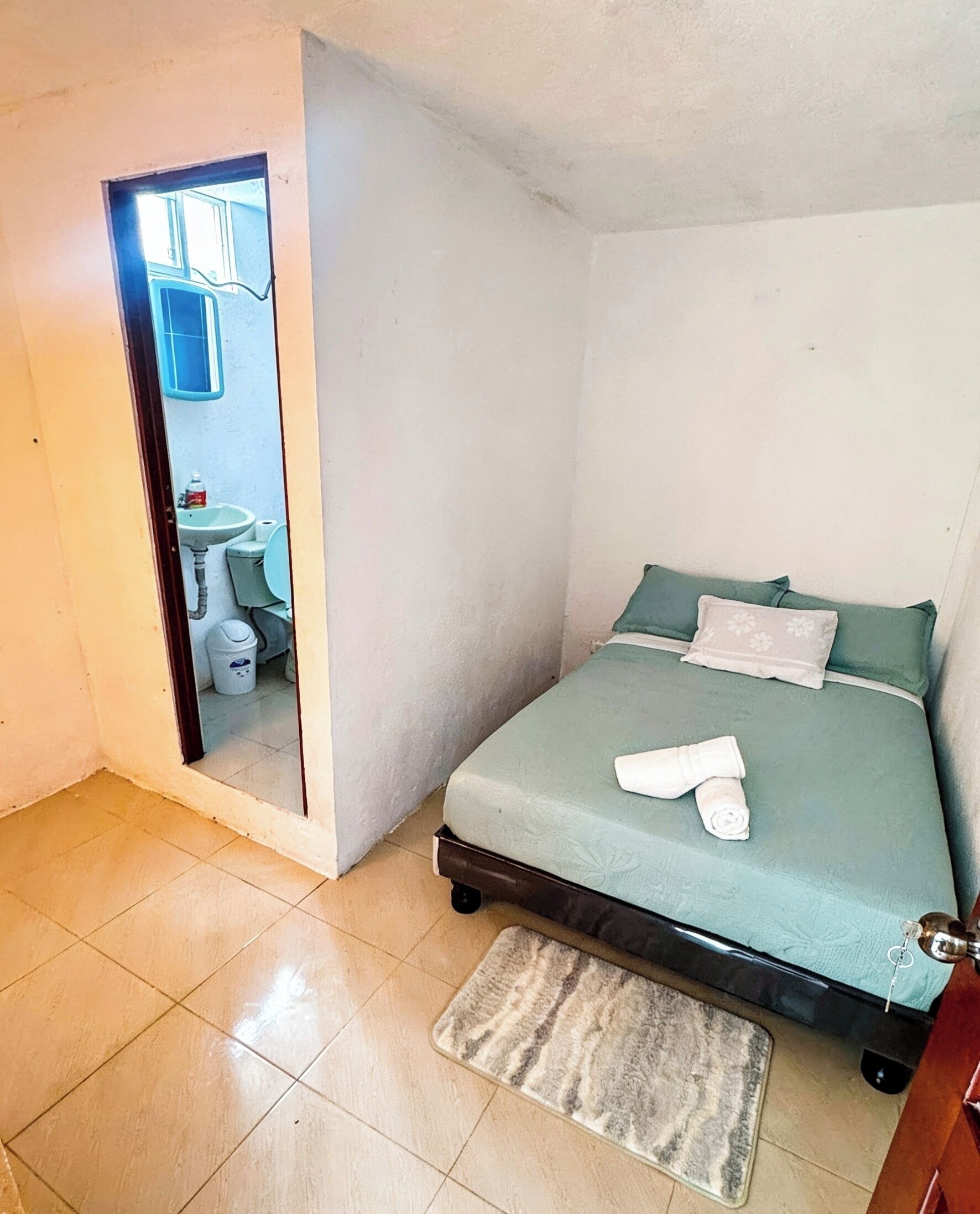 Una camera piccola con un letto singolo, materasso verde, porta in legno, pavimento piastrellato e bagno con lavandino e WC.