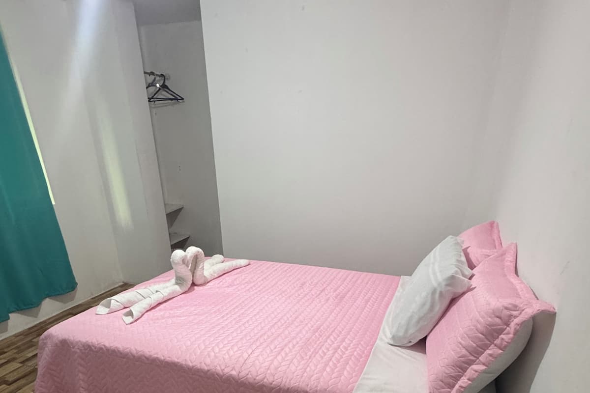 Un lit bien fait, avec une couvre-lit rose et des oreillers blancs, une table de chevet en bois et un porte-manteau fixé au mur.