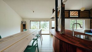 Een houten eettafel met stoelen, een keukenwerkblad met een spoelbak en grote ramen.