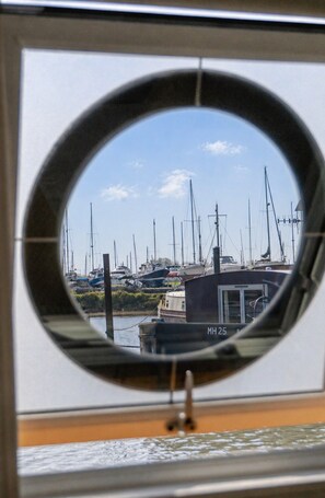 Una ventana circular con vista a un puerto deportivo, barcos y un edificio con el identificador 'MH 25'.