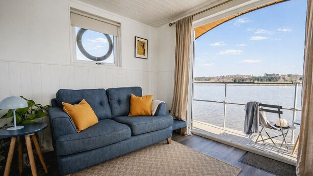 A living room with a blue sofa, a small side table, a potted plant, a window with a view of a body of water, and a door leading to an outdoor area.
