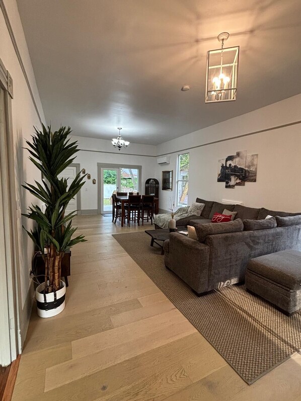 A spacious living room with a grey sectional sofa, a dining area with a table and chairs, and a large potted plant.
