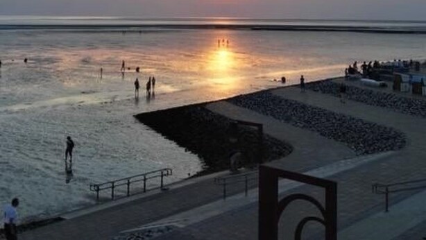 Ein Sonnenuntergang über einem Strand, mit Menschen, die am Ufer entlanggehen, und einem Promenadenweg.