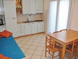 A compact kitchen with white cabinets, a gas stove, and a sink. There is a dining table with chairs and a blue sofa with an orange pillow. - Wohnung im Zentrum von Lignano Sabb. "Maria 1 by Interhome (Lignano Pineta)
