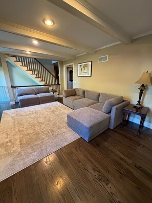 A living room with a large sectional sofa, a small ottoman, a wooden floor, a staircase, and a lamp. - Spacious 6-bedroom house in KC suburb. 20 minutes from Arrowhead Stadium. (Leawood)
