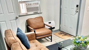 A living room with a brown sofa, a brown armchair, a glass coffee table, and a door with a doormat.