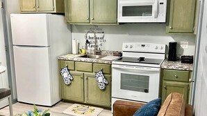 A compact kitchen with green cabinets, a white refrigerator, a microwave, and a stove. There is a glass coffee table and a brown sofa with blue cushions.