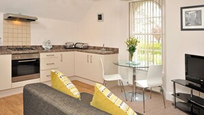 Een moderne keuken met een gasfornuis, oven en een ingebouwde magnetron. Er is een eethoek met een glazen tafel en witte stoelen. Een televisie staat op een zwarte standaard.