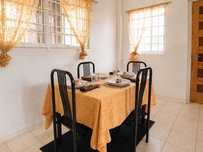 A dining area with a table set for four, black chairs, a yellow tablecloth, and lace curtains. - Lucian Home. Fish Friday Village. Full House (Anse La Raye)
