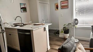 A compact kitchen with a sink, a dining area with a table and chairs, a grey sofa, and a potted plant. - Lib3rtie Suites -2BR Family Suite •Sleeps 6 •Full Kitchen •Laundry •Free Parking (Saskatoon)