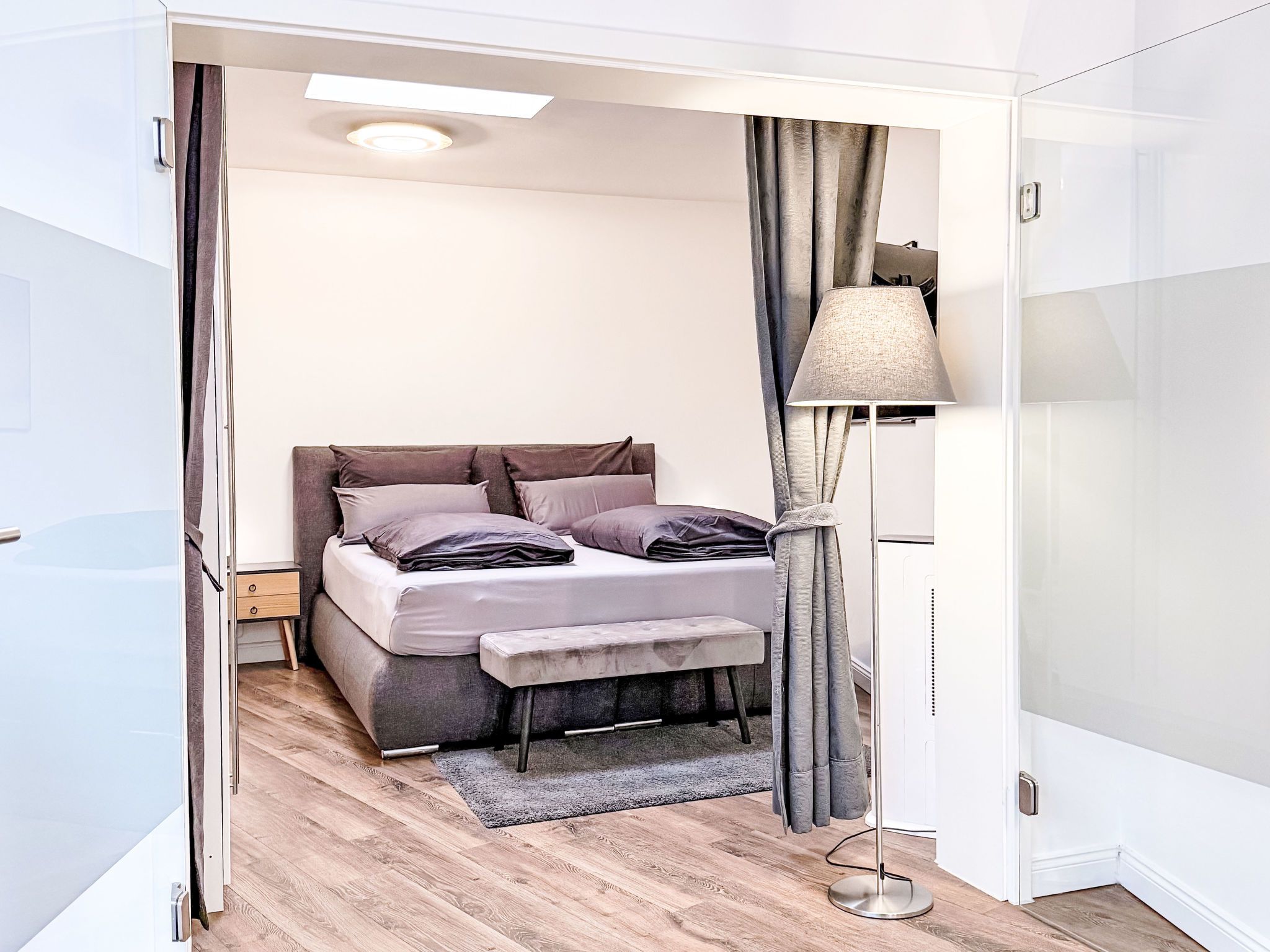 Ein Schlafzimmer mit Bett, Nachttisch, Lampe und Blick durch eine offene Tür.