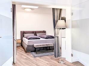 Une chambre à coucher avec un lit, une table de chevet, une lampe et une vue sur l’extérieur, grâce à une porte ouverte.