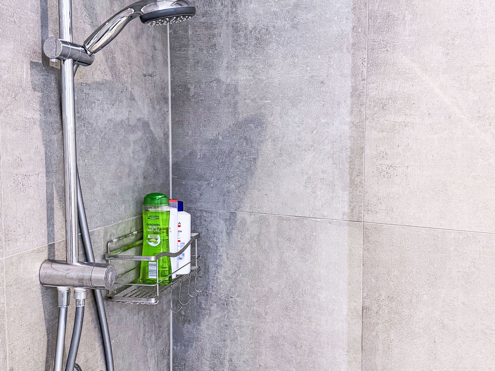 A shower area with a metal faucet and a water stream, a metal shelf with bottles, and a textured wall.
