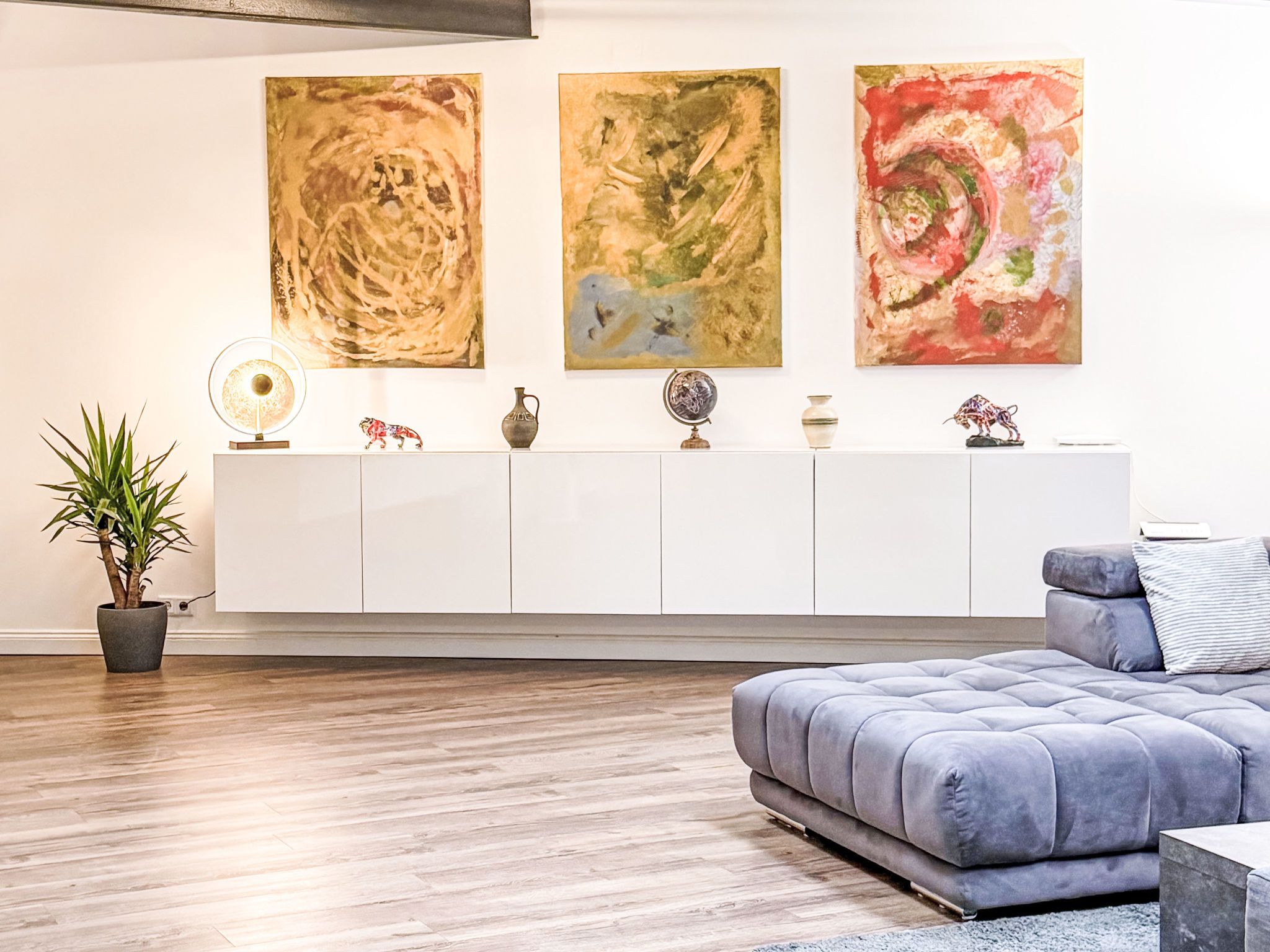 Una sala de estar moderna con un sofá gris, una mesa consola blanca con objetos decorativos y pinturas abstractas en la pared.