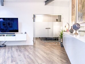 A modern living room with a flat-screen TV, a white entertainment unit, a glass coffee table, and a potted plant.