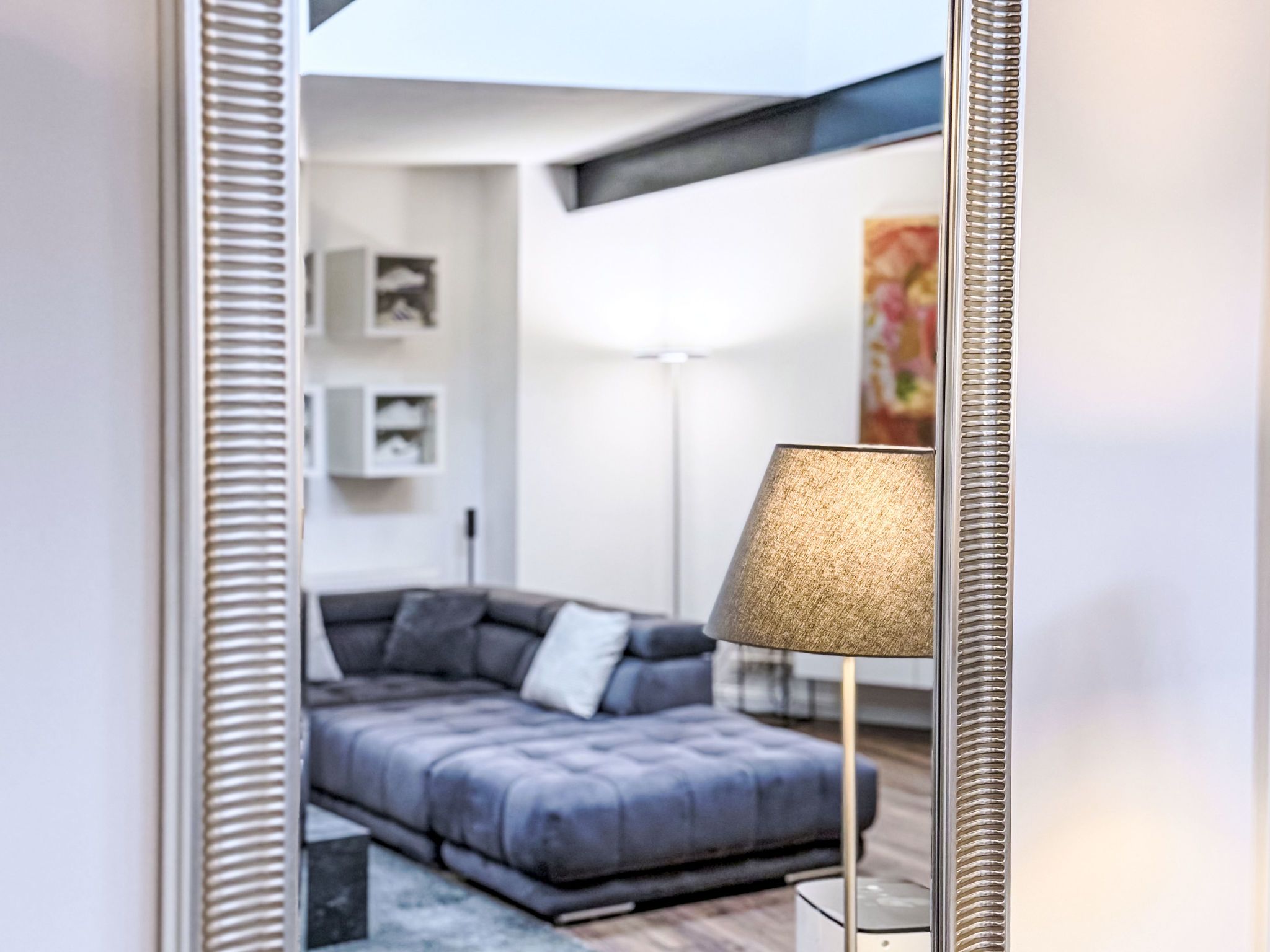 Ett modernt vardagsrum med en grå soffa, en golvlampa och en spegel som reflekterar rummet.