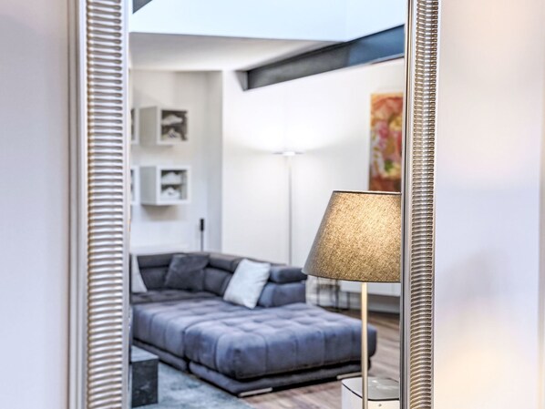 A modern living room with a grey sofa, a floor lamp, and a mirror reflecting the room.
