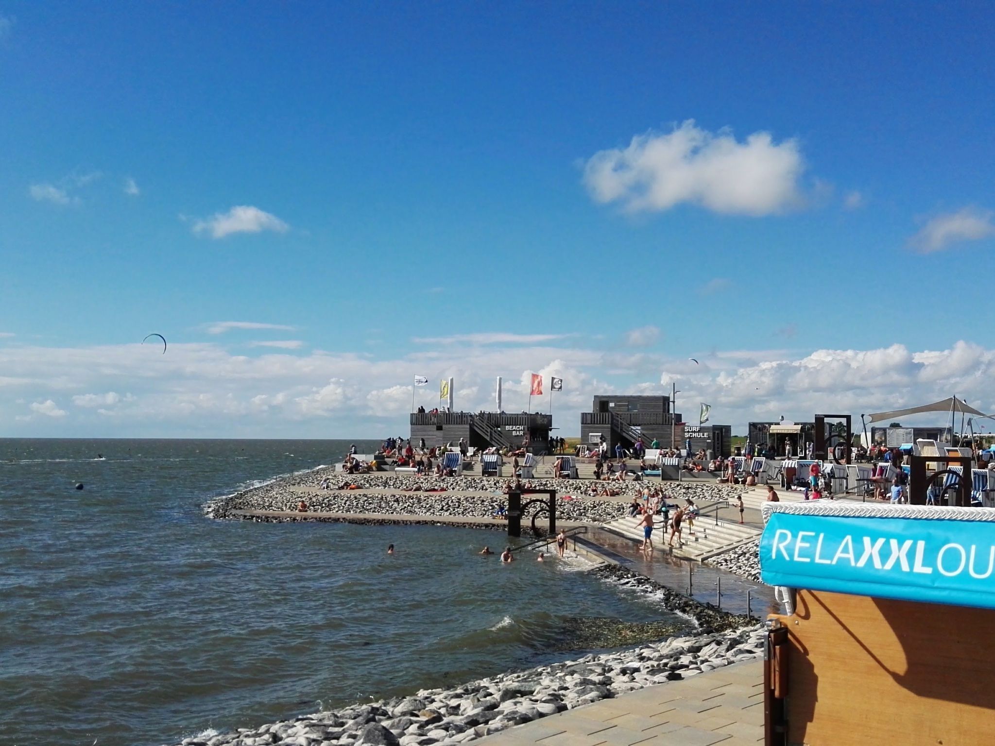 Ubicación frente a la playa con un muelle de madera, un bar llamado 'BEACH BAR' y un cartel de 'RELAXXLOU'.