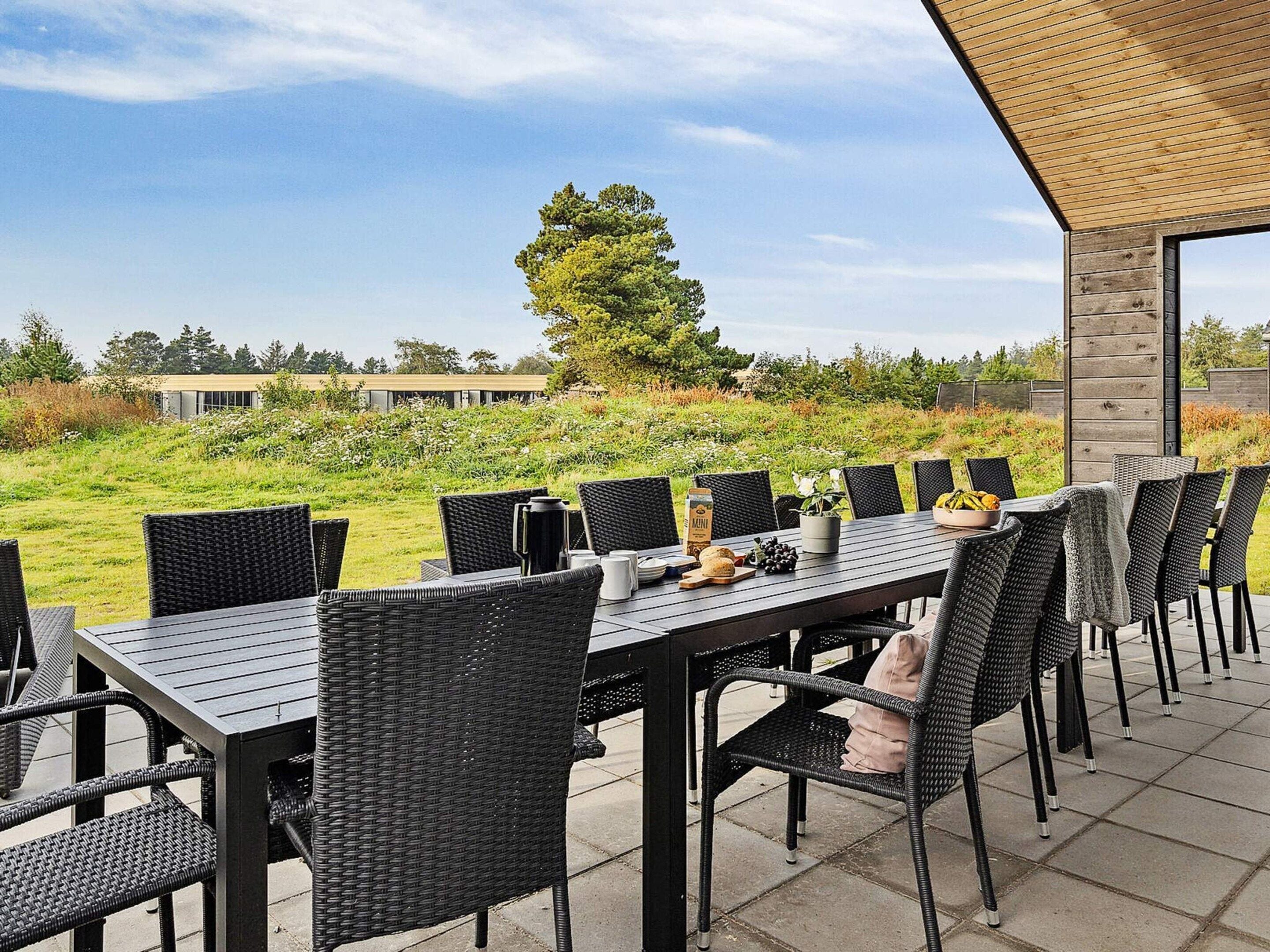 Een terras met een lange tafel en stoelen, omgeven door een gazon en een gebouw op de achtergrond.
